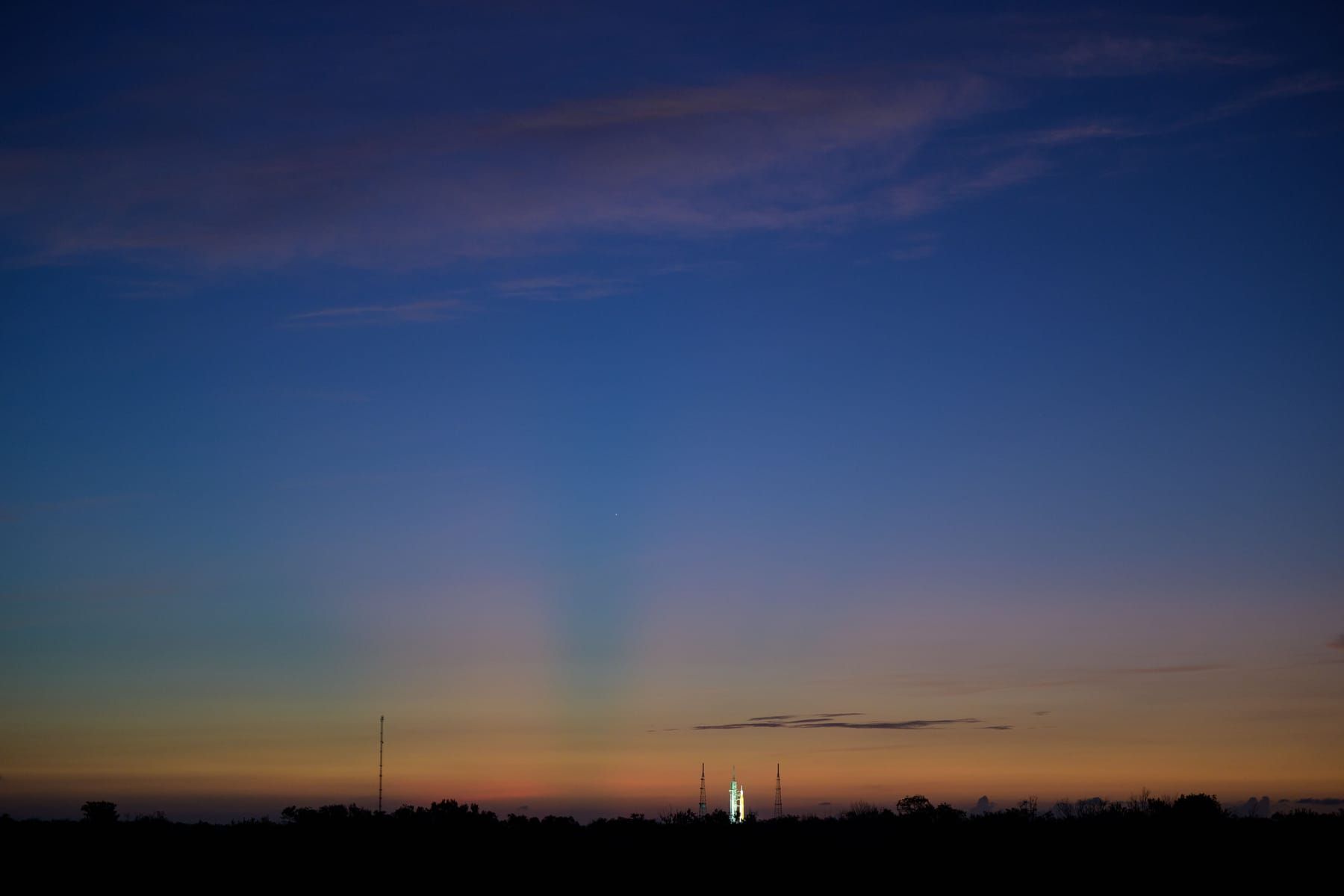 Deep blue sky fading to orange at the horizon, with a lit-up rocket on its launch pad in the distance