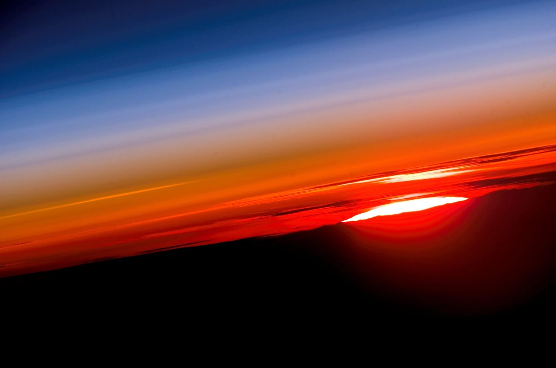 Setting Sun squashed into a flat, bright oval at Earth's horizon beneath red, orange and blue layers of atmosphere