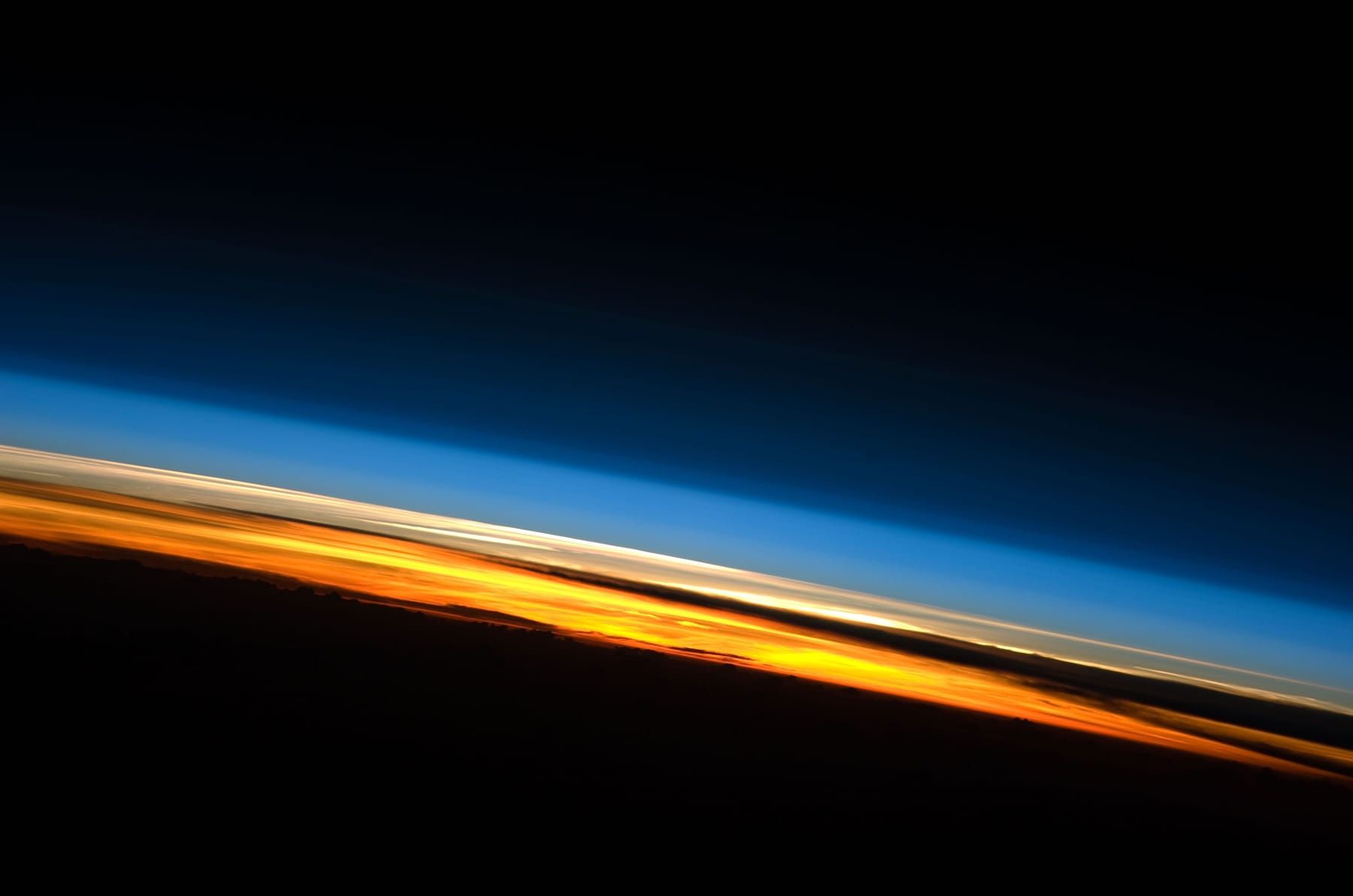 Thin curved band of Earth's atmosphere at sunset seen from orbit, layered orange, white and blue above the dark surface